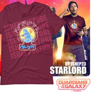 May include: A maroon t-shirt with a graphic of a blue dinosaur holding a blue orb. The graphic is surrounded by the text "JSHEP23STORE" in a white, blue, and yellow color scheme. The text "BY JSHEP23 STARLORD" is printed below the graphic. The text "GUARDIANS OF THE GALAXY VOLUME 3" is printed below the text "STARLORD".
