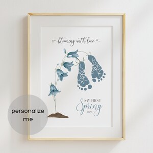 May include: Framed art print with a light wood frame. The print features bluebell flowers and two blue footprints. Text reads "blooming with love" and "My First Spring 2026". A circular graphic says "personalize me".