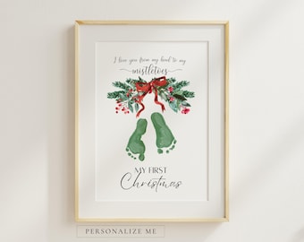May include: A framed print featuring a watercolour illustration of green baby footprints, a pine branch with red berries, and a red bow. The text reads: I love you from my head to my mistletoes, and My First Christmas. The frame is light wood.