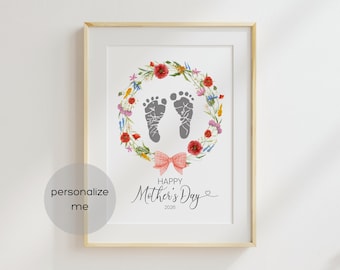 May include: Framed print with a floral wreath surrounding two gray baby footprints. A pink bow sits below the wreath, with the words "Happy Mother's Day 2026" in elegant script. The print is in a light wood frame.