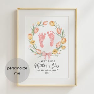 Op de afbeelding: Ingelijste print met een licht houten frame. Het kunstwerk toont een bloemenkrans met roze en gele tulpen, rondom roze babyvoetjes. De tekst luidt "Happy First Mother's Day as my Grandma 2026". Onderaan een roze strik.