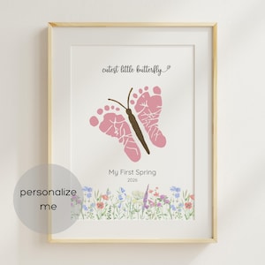 May include: Framed print featuring a pink butterfly made from baby footprints. The print includes the text "cutest little butterfly" and "My First Spring 2026". A floral border decorates the bottom of the print.