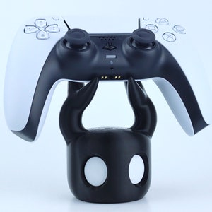 Pode incluir: Um suporte para controlador de videogame preto e branco em forma de personagem do videogame Hollow Knight. O suporte está segurando um controlador PlayStation 5 branco e preto.