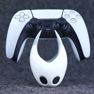 May include: A white PlayStation 5 DualSense controller with black accents is placed on top of a white 3D printed figure of the Knight from the video game Hollow Knight.