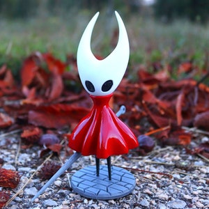 May include: A white and red figurine of a character from the video game Hollow Knight. The character is wearing a red dress and has large white horns. The figurine is standing on a gray base with a sword.