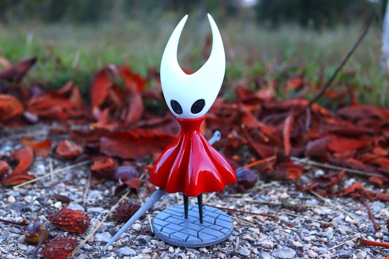 May include: A white and red figurine of a character from the video game Hollow Knight. The character is wearing a red dress and has large white horns. The figurine is standing on a gray base with a sword.