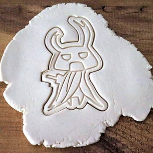 May include: A white cookie cutter in the shape of a cartoon character, pressed into a piece of dough. The character has a horned head, a face with two eyes, and a long, flowing garment.