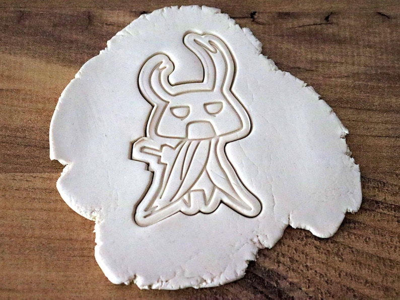May include: A white cookie cutter in the shape of a cartoon character, pressed into a piece of dough. The character has a horned head, a face with two eyes, and a long, flowing garment.