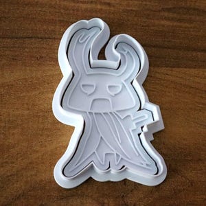 May include: A white plastic cookie cutter in the shape of a stylized character with a horned head, a cape-like design, and a stern expression. The cutter is on a brown wooden surface.