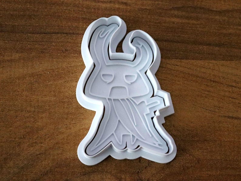 May include: A white plastic cookie cutter in the shape of a stylized character with a horned head, a cape-like design, and a stern expression. The cutter is on a brown wooden surface.