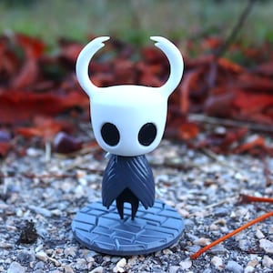 May include: A white and gray figurine of a character from the video game Hollow Knight. The figurine is standing on a gray base with a cracked pattern. The character has a white mask with two black eyes and two long, pointed horns.