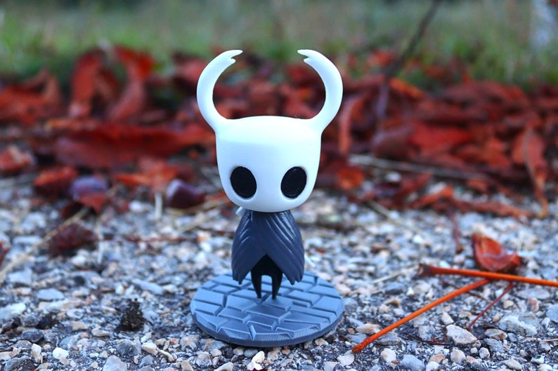 May include: A white and gray figurine of a character from the video game Hollow Knight. The figurine is standing on a gray base with a cracked pattern. The character has a white mask with two black eyes and two long, pointed horns.