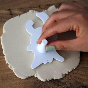 May include: A white bunny-shaped cookie cutter being pressed into a piece of light-colored dough. The cutter is held by a hand with painted nails, creating a bunny shape. The dough sits on a wooden surface, suggesting a baking or crafting activity.