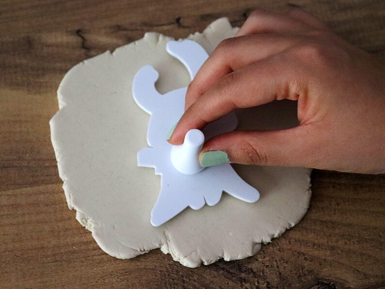 May include: A white bunny-shaped cookie cutter being pressed into a piece of light-colored dough. The cutter is held by a hand with painted nails, creating a bunny shape. The dough sits on a wooden surface, suggesting a baking or crafting activity.