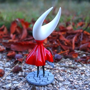 May include: A white and red figurine of a character from the video game Hollow Knight. The character is standing on a gray base with a cobblestone design. The figurine is posed with its arms outstretched and its head tilted back.