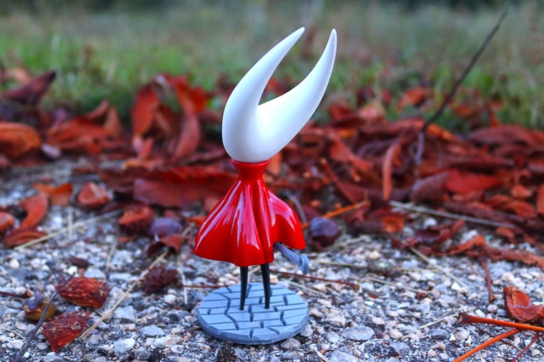 May include: A white and red figurine of a character from the video game Hollow Knight. The character is standing on a gray base with a cobblestone design. The figurine is posed with its arms outstretched and its head tilted back.