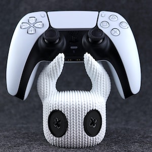 Könnte beinhalten: Ein weißer, gestrickter Controller-Ständer in Form einer Kreatur mit zwei großen Ohren und zwei schwarzen Knöpfen für die Augen. Ein weißer PlayStation 5-Controller liegt auf dem Ständer.