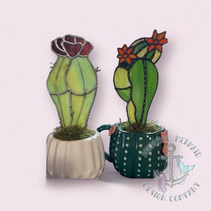 Op de afbeelding: Twee glas-in-lood cactusplanten in keramische potten. De cactus links is groen met een rode bloem en de cactus rechts is groen met oranje bloemen. De potten zijn beide versierd met stippen. De pot rechts is groen en de pot links is wit. De tekst "Anchored Mermaid Design Company" is zichtbaar aan de rechterkant van de afbeelding.