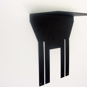 May include: A modern, black metal wall shelf with a geometric design. The shelf has a rectangular top and a unique, angular support structure with vertical cutouts. The shelf is mounted on a white wall.