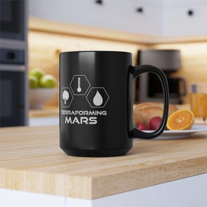 May include: A black ceramic mug featuring a white graphic with three hexagonal icons representing a tree, a thermometer, and a water droplet. Below the icons, the text reads TERRAFORMING MARS in a bold, clean, sans-serif font style.