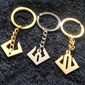 May include: Three keychains with geometric pendants. Two are gold-toned, and one is silver-toned. Each keychain has a ring and a chain connecting to a square pendant with a unique cutout design. The keychains are displayed on a dark, textured surface.