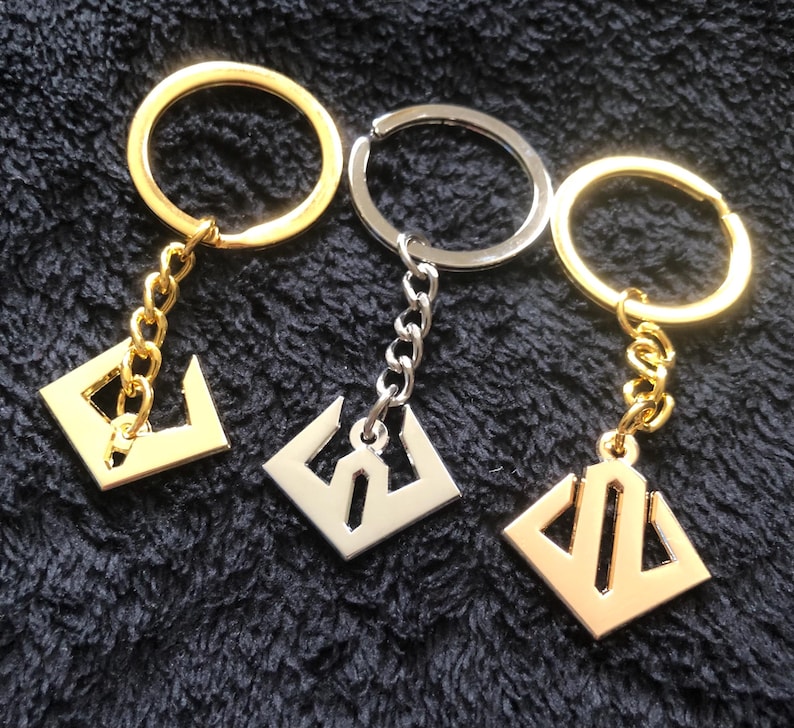 May include: Three keychains with geometric pendants. Two are gold-toned, and one is silver-toned. Each keychain has a ring and a chain connecting to a square pendant with a unique cutout design. The keychains are displayed on a dark, textured surface.