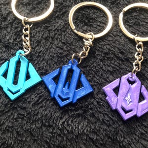 May include: Three keychains with geometric pendants in light blue, dark blue, and purple. Each pendant is attached to a silver chain and a silver key ring. The keychains are displayed on a dark, textured surface.