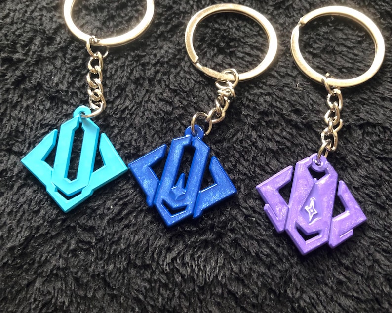 May include: Three keychains with geometric pendants in light blue, dark blue, and purple. Each pendant is attached to a silver chain and a silver key ring. The keychains are displayed on a dark, textured surface.