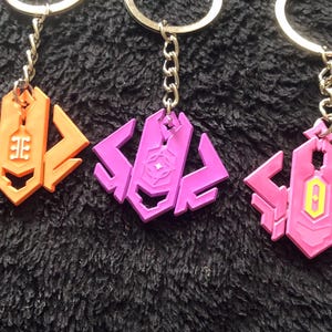 May include: Three keychains with geometric designs in orange, purple, and pink. Each keychain has a silver chain and ring. The orange keychain has a white symbol, the purple has a star, and the pink has a yellow and white symbol.