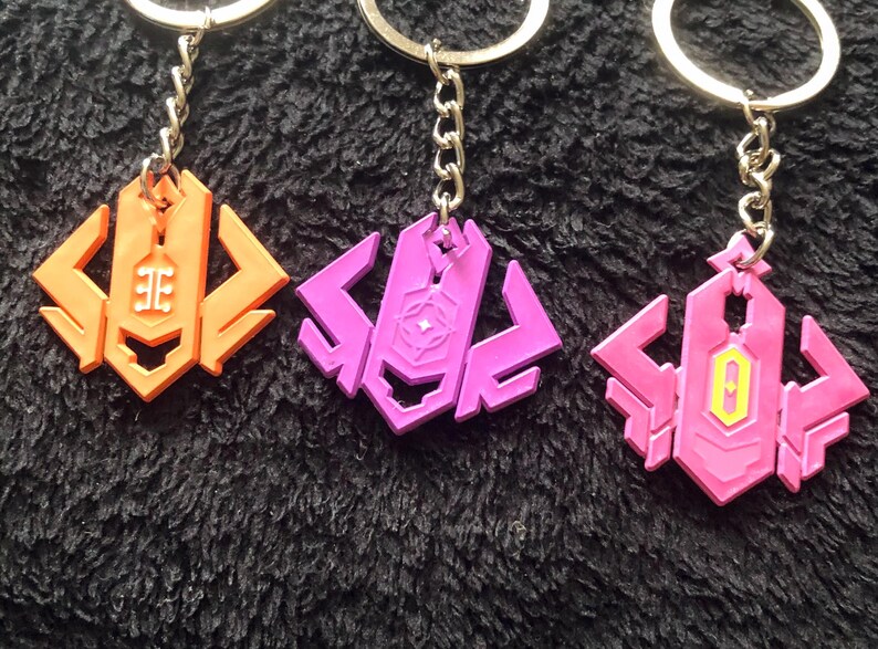 May include: Three keychains with geometric designs in orange, purple, and pink. Each keychain has a silver chain and ring. The orange keychain has a white symbol, the purple has a star, and the pink has a yellow and white symbol.
