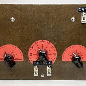Könnte beinhalten: Ein Vintage-Radioempfänger mit einer braunen Holzfrontplatte. Es verfügt über drei rote Zifferblätter mit schwarzen Knöpfen, die mit "PHONES", "ANT" und "CRYSTAL" beschriftet sind. Die Zifferblätter haben Zahlen von 0 bis 7. Das Design ist retro.