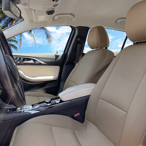 May include: Beige leather car seats with headrests in a car interior. The seats are in the front passenger and driver positions. The car has a black steering wheel and a black center console.