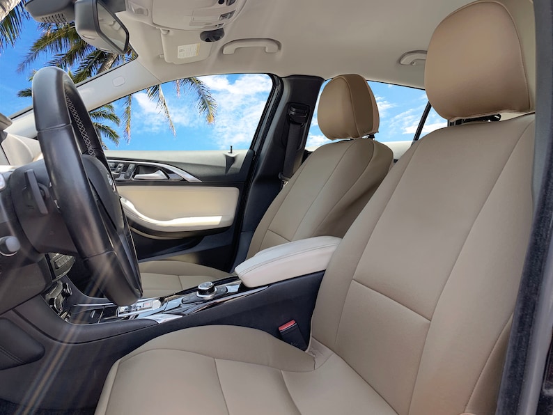 May include: Beige leather car seats with headrests in a car interior. The seats are in the front passenger and driver positions. The car has a black steering wheel and a black center console.