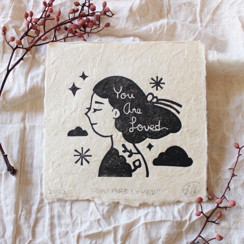 A girl looking to the left with her eyes closed and the words "You Are Loved" etched into her hair. She is surrounded by clouds and stars.