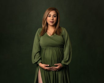May include: A pregnant woman in a long, green, flowing dress with a slit up the side. She is standing in front of a dark green background and is holding her belly.