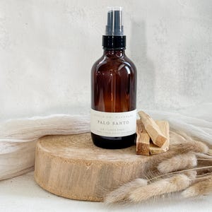 May include: A brown glass bottle with a black spray nozzle and a white label that reads "PALO SANTO AIR + LINEN SPRAY." The bottle sits on a wooden disc with several pieces of Palo Santo wood next to it. Dried plants are in the foreground.