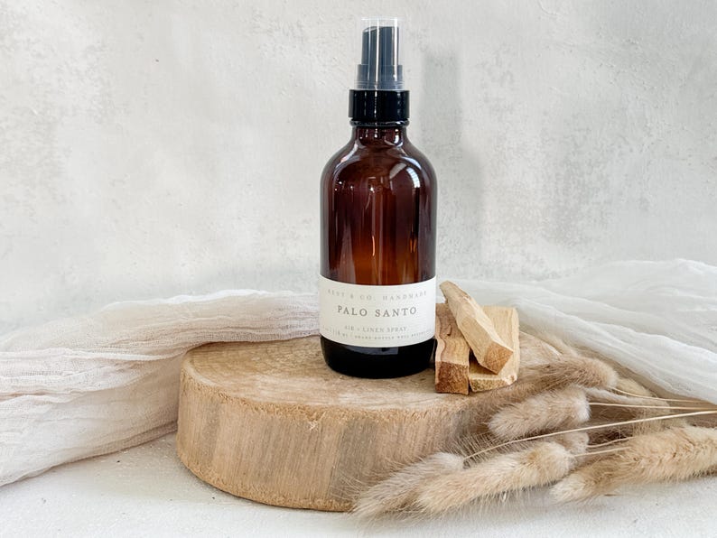 May include: A brown glass bottle with a black spray nozzle and a white label that reads "PALO SANTO AIR + LINEN SPRAY." The bottle sits on a wooden disc with several pieces of Palo Santo wood next to it. Dried plants are in the foreground.