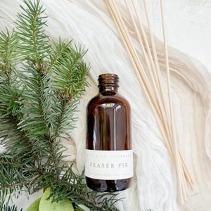 May include: A brown glass bottle with a white label that reads "Fraser Fir Reed Diffuser 1.8 oz/53 ml" and a set of wooden diffuser sticks. The bottle is surrounded by green pine branches and leaves.