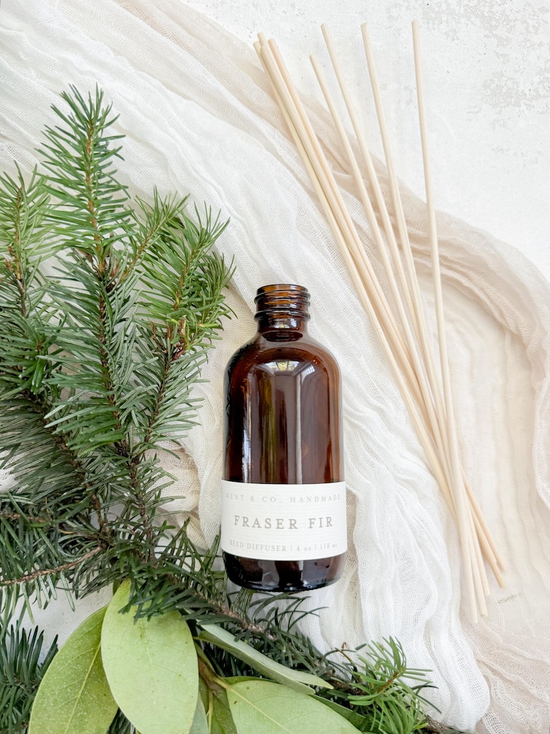 May include: A brown glass bottle with a white label that reads "Fraser Fir Reed Diffuser 1.8 oz/53 ml" and a set of wooden diffuser sticks. The bottle is surrounded by green pine branches and leaves.