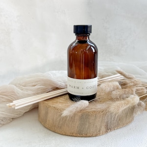 May include: A brown glass bottle with a black lid and a white label that reads "Warm + Cozy Reed Diffuser" on a wooden surface with dried grass.