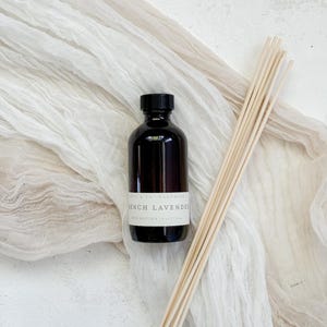 May include: A dark brown glass bottle with a black lid and a white label that reads "French Lavender Reed Diffuser 1 oz/30 ml". The bottle is sitting on a white surface with a bundle of natural wooden sticks.