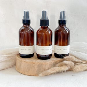 May include: Three amber glass spray bottles with black spray tops and white labels, each containing a different scent. The bottles are displayed on a wooden disc with dried plants and a white cloth in the background. The labels read "Palo Santo" and "Macintosh Apple".