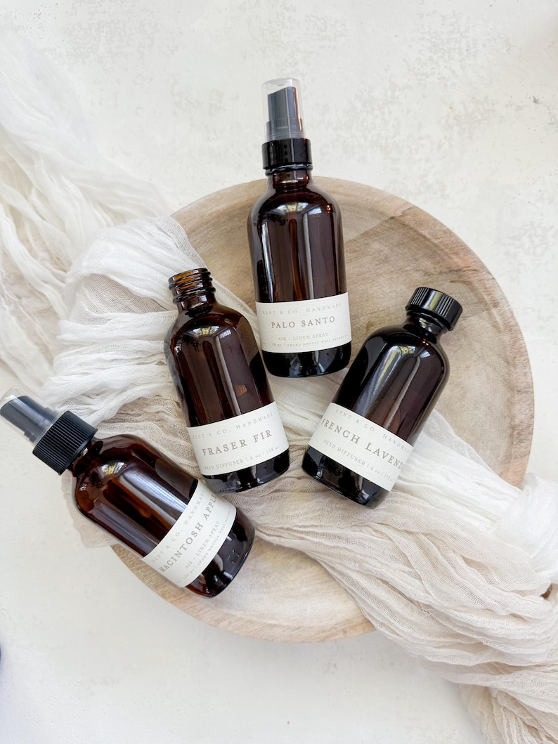 May include: Four amber glass bottles with black lids and white labels containing room spray. The labels have the text "Mist & Co. Handmade" and the scent names "Palo Santo", "Fraser Fir", "French Lavender", and "Macintosh Apple". The bottles are arranged on a wooden tray with a white fabric background.