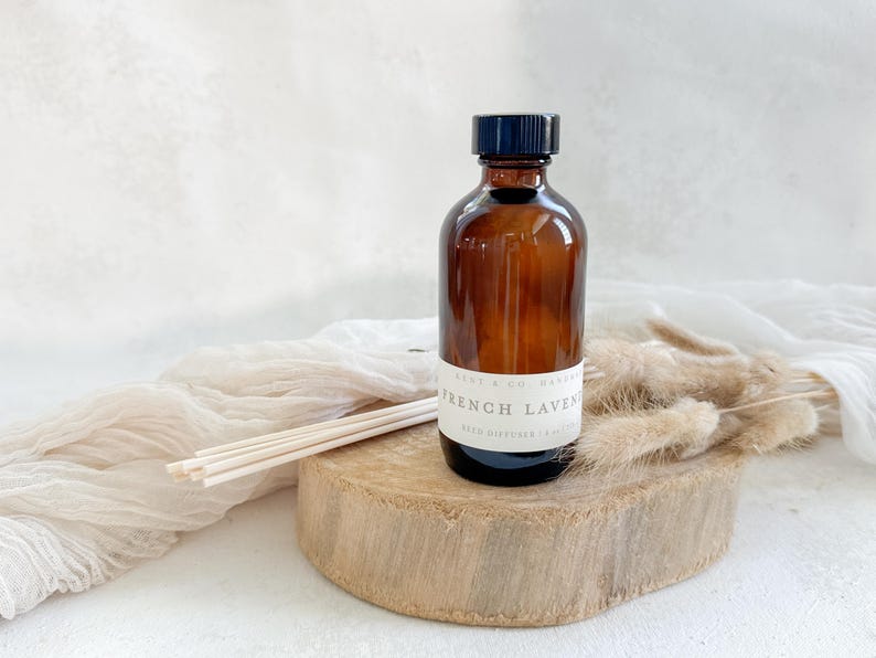 May include: A brown glass bottle with a black lid and a white label that reads "French Lavender Reed Diffuser 100ml". The bottle is sitting on a wooden surface with dried grass and a white fabric in the background.
