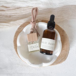 May include: Two small glass bottles with brown liquid and labels. The bottle on the left is a car diffuser refill with the word "Hand" written on the label. The bottle on the right is a car diffuser refill with the text "Palo Santo Car Diffuser Refill Small Batch Made in WA". The bottles are sitting on a white marble dish with a wooden base.
