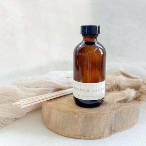 May include: A brown glass bottle with a black lid and a white label that reads "French Lavender Reed Diffuser 100ml". The bottle is sitting on a wooden surface with some dried grass.