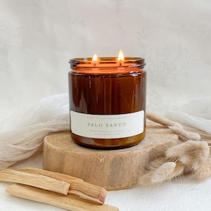 May include: A brown glass candle jar with two lit wicks. The candle is labeled "Palo Santo" and is sitting on a wooden surface with dried grass.
