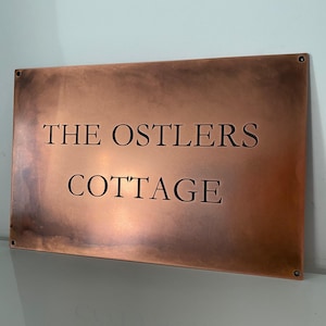 Op de afbeelding: Een rechthoekig koperen bord met de woorden "THE OSTLERS COTTAGE" in een schreefletter. Het bord heeft een warme, roodbruine tint en is op een witte muur gemonteerd. Het bord is waarschijnlijk voor een huisje of huis.