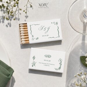 May include: Two white matchboxes with green floral and text details. One box is open, revealing matches. The text on the boxes includes initials, a date, and location. The image is styled with a white plate, a green napkin, and floral accents.