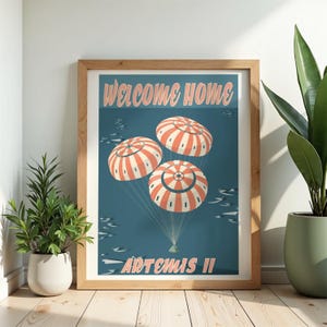 May include: Wooden framed art print with a retro illustration of three parachutes descending on a teal background. The text "WELCOME HOME" and "ARTEMIS II" are displayed in a vintage font.